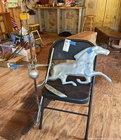 Metal weather vane with running horse figure positioned on a black folding chair indoors.