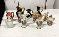 Front view of 11 miniature Limoges urns and jugs, showing various colors and hand-painted courting couple designs, predominantly white with gold and some with blue and pink coloring.
