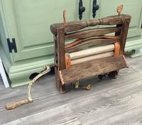 Photo showing antique clothing wringer with wooden frame, cloth rollers, and cast iron handle resting on floor near light colored cabinet.