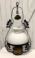 Antique hanging kerosene lamp showing solid white glass shade, black metal decorative frame with scrollwork, and brass fuel container at the base.
