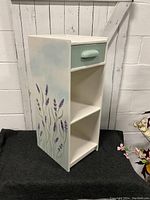 Full view of the side table showing white paint, hand painted lavender flowers on the side, and the soft green drawer front with rounded handle.