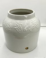 Front and side view of large cream-colored crock showing raised grape pattern and pre-drilled hole near the base with tape