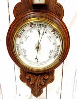 Close-up of barometer dial showing pressure readings and weather conditions, mounted in wooden carved frame with brass bezel.