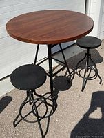 Full view of the bistro set with round solid Acacia wood table top and two round black adjustable metal stools.