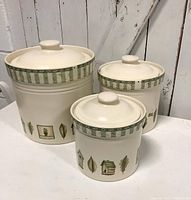 Three cream-colored stoneware canisters with lids, showing full set and design details with nature-themed decals and green rim pattern.