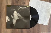 Photo of the LP cover, vinyl disc, and lyric sheet laid out on floor. Shows black and white photo on cover of John Lennon and Yoko Ono, with yellow text.