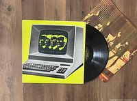 Photo showing the Kraftwerk Computer World LP vinyl record partially out of its yellow-bordered album cover with retro computer and band images, alongside the inner sleeve featuring band photos.