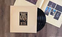 Three black vinyl records partly pulled out of a cream colored gatefold sleeve with gold and black text The Band The Last Waltz. Open booklet with photos of band members and concert inside.