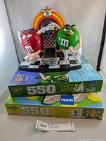 Front view of M&Ms candy dispenser featuring red and green characters in front of a jukebox on top of two puzzle boxes.