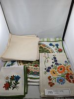 Grouped vintage linens showing colorful hand embroidered floral patterns on linen cloths with some pieces folded showing embroidered details and plain linen napkins.