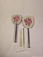 Front view of vintage handheld mirror and hairbrush showing pink floral patterned backs, with small white comb in center.