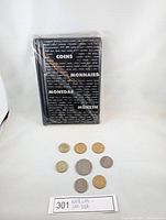 Sealed black coin book and nine assorted coins arranged in front on white background with lot number.