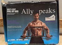 Box with branding for Ally Peaks multi-grip pull up bar, showing male model demonstrating use with multiple grip options. Packaging is black with white and blue text.