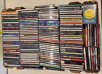 Photo showing a large box of stacked used CDs, some with cracked cases, from various artists and genres.