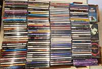 Photo showing a large assortment of used music CDs in various jewel cases stacked in rows within a cardboard box, also visible are a few cassette tapes on the side.