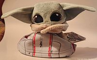Close-up image of The Child plush with large eyes and beige cloak, sitting inside a gray and red hover-pram ball