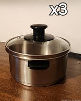 Three stainless steel steamers each with a glass lid and black handle, side grips visible, placed on wooden surface, photo shows a front view.