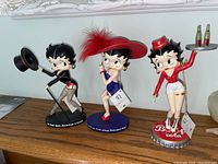 Three Betty Boop figurines displayed on a shelf: Carhop Berry with soda tray, Its Not the Age Betty with blue dress and red feather hat, and Dancer Betty in black costume holding top hat