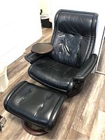 Full view of black leather lounge chair with attached round wood side table and matching ottoman on wood floor