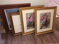 Four framed artworks leaning against a wall on the floor, showing varying frame styles and art subjects.