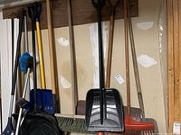 Photo showing assorted lawn and garden tools including rakes, shovels, and brooms standing against a wall