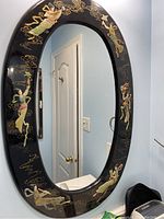 Full view of oval wall mirror showing black lacquered frame with multiple painted Asian female figures in traditional attire around the edge, with background showing room interior including door and light switch.