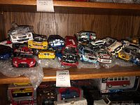 Multiple racing model cars on wooden shelf with bubble wrap and some with visible wear and missing parts
