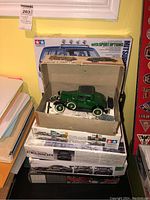 Open beige box with assembled green 1930s coupe model, loose parts and instructions visible; stack of model kit boxes and Tamiya Montero kit in background
