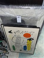 Two framed abstract art prints leaning against a wall wrapped partially in bubble wrap. One print prominently displays an orange circle and black star shape with hand-drawn text including 'Miro' and 'Maegh'.