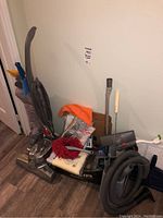 Gray Sedition upright vacuum cleaner with coiled power cord and multiple vacuum accessories, cleaning cloths, and parts gathered together against a wall on floor.
