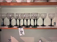Shelf holding 10 clear wine glasses with black stems and bases, arranged in a row.