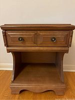 Front view of wooden end table showing drawer front, lower shelf, decorative edges, and floor placement.