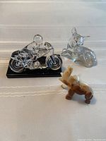 Three figurines displayed on a white cloth background: a clear crystal motorcycle and rider on black base, a glass fawn, and a marble elephant.