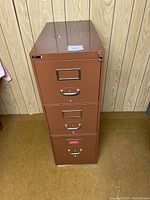 Closed view of the brown metal filing cabinet showing three drawers with pull handles and label holders.
