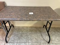 Front view of the folding table showing brown marbled laminate top and black metal legs on tiled floor.