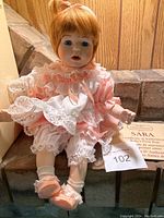 Porcelain doll named Sara with blond hair, blue eyes, wearing a pink and white lace dress, with Certificate of Authenticity displayed beside the doll