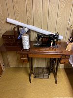 Singer treadle sewing machine mounted on wooden table with drawers and treadle base, with large spool of white thread and rolled white fabric on top.