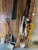 Photo of various garden tools including shovels, broom, dustpan, and a yellow electrical cord hanging on a wood panel wall.