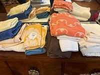 Photo showing assorted towels folded and stacked on a table, with variety of colors and patterns visible including yellow vintage pattern and blue solid towel.