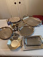 Photo showing multiple serving trays arranged on table with various shapes and decorative edges. Included are two metal goblets.