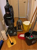 Overall view of the Fantom and Shark upright vacuum cleaners, yellow bucket with lid, wooden mop handle, dustpan with brush, and black wringer bucket with cleaning supplies on hardwood floor.