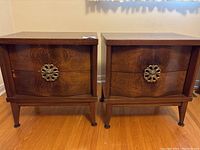Front view of two vintage wooden Art Deco end tables with metal flower-shaped drawer pulls, set on hardwood floor.