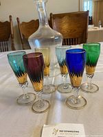 Set of six vintage champagne glasses with gold accents and various colors arranged around a clear glass decanter on a white tablecloth