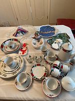 Top view of vintage Christmas-themed china and glassware arranged on a white tablecloth. Items include cups, saucers, plates, creamer, sugar bowl, and decorative holiday figurines including snowmen and tree shapes.