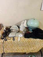 Photo of vintage hairdresser items on yellow floral tablecloth including Lady Schick hood dryer, curling irons, hair clipper, handheld mirror, cape, and various accessories.