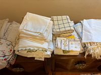 Photo shows a collection of folded white tablecloths, beige and blue checkered linens, and white fabric with fringed edge, placed on a wooden surface.
