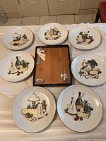 Six vintage white serving plates with various cheese and food patterns arranged around a wooden cheese board with metal utensils.