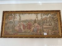 Full view of large rectangular tapestry showing detailed scene with 18th century men and women in period dress, set in outdoor scenic garden with classical architecture and water body in background. Mounted in an ornamental gilt frame.
