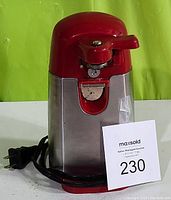 Front view of AmazonBasics red electric can opener showing red upper housing, stainless steel body, manual lever, cutting blade, power cord, and plug.