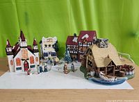 Wide front view of four Christmas village buildings with figurines arranged on white surface against green background.